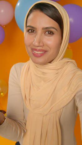 Retrato-De-Estudio-De-Video-Vertical-De-Una-Mujer-Que-Usa-Hijab-Celebrando-Un-Cumpleaños-Soplando-Confeti-De-Papel-Rodeado-De-Globos